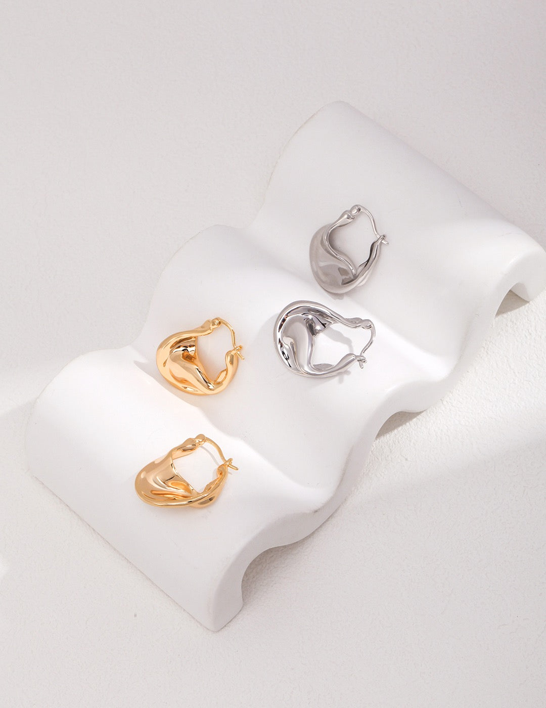 Organic fluid hoop earrings in 18K gold and 18K white gold plating, made from 925 sterling silver, displayed on a white curved stand