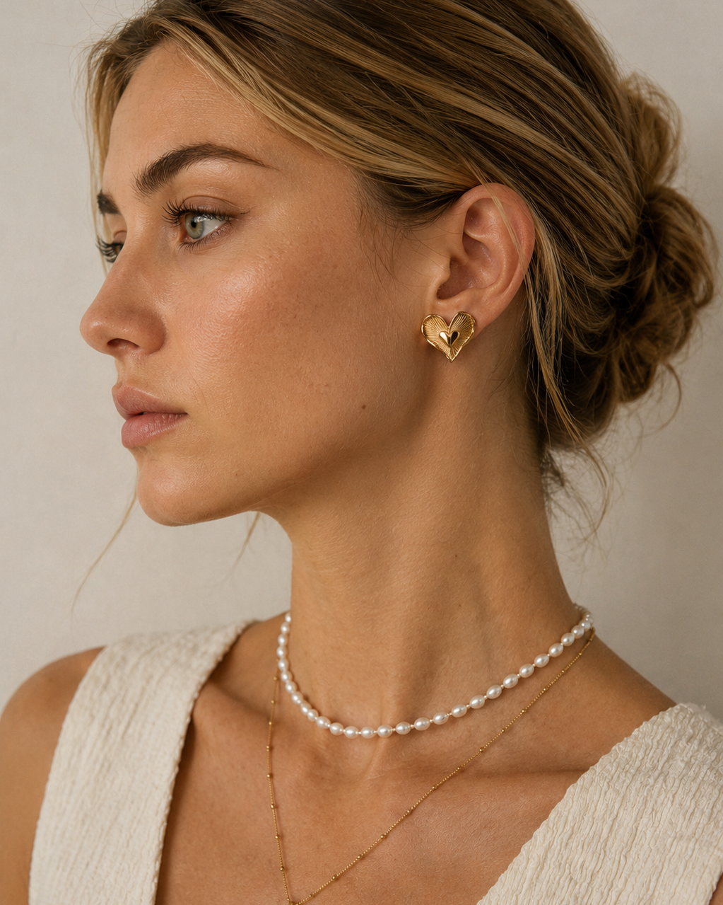European model wearing Amour Radiance heart stud earrings in thick 18K gold plating, styled with layered necklaces in an elegant side-profile portrait.
