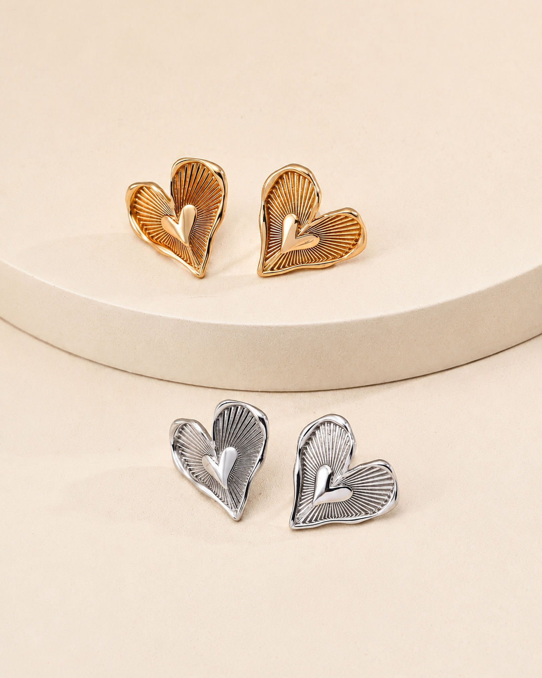 Amour Radiance heart stud earrings displayed in both thick 18K gold plated and thick 18K white gold plated finishes on a minimalist cream pedestal.