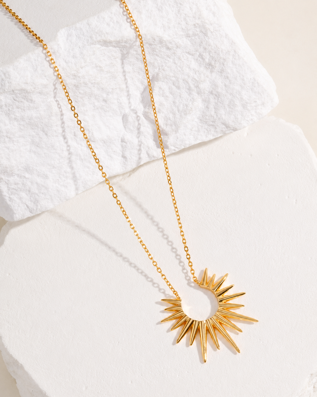 Gold sunburst pendant necklace displayed on a white textured stone slab against a soft beige background, featuring a radiant sun-inspired design and delicate chain.