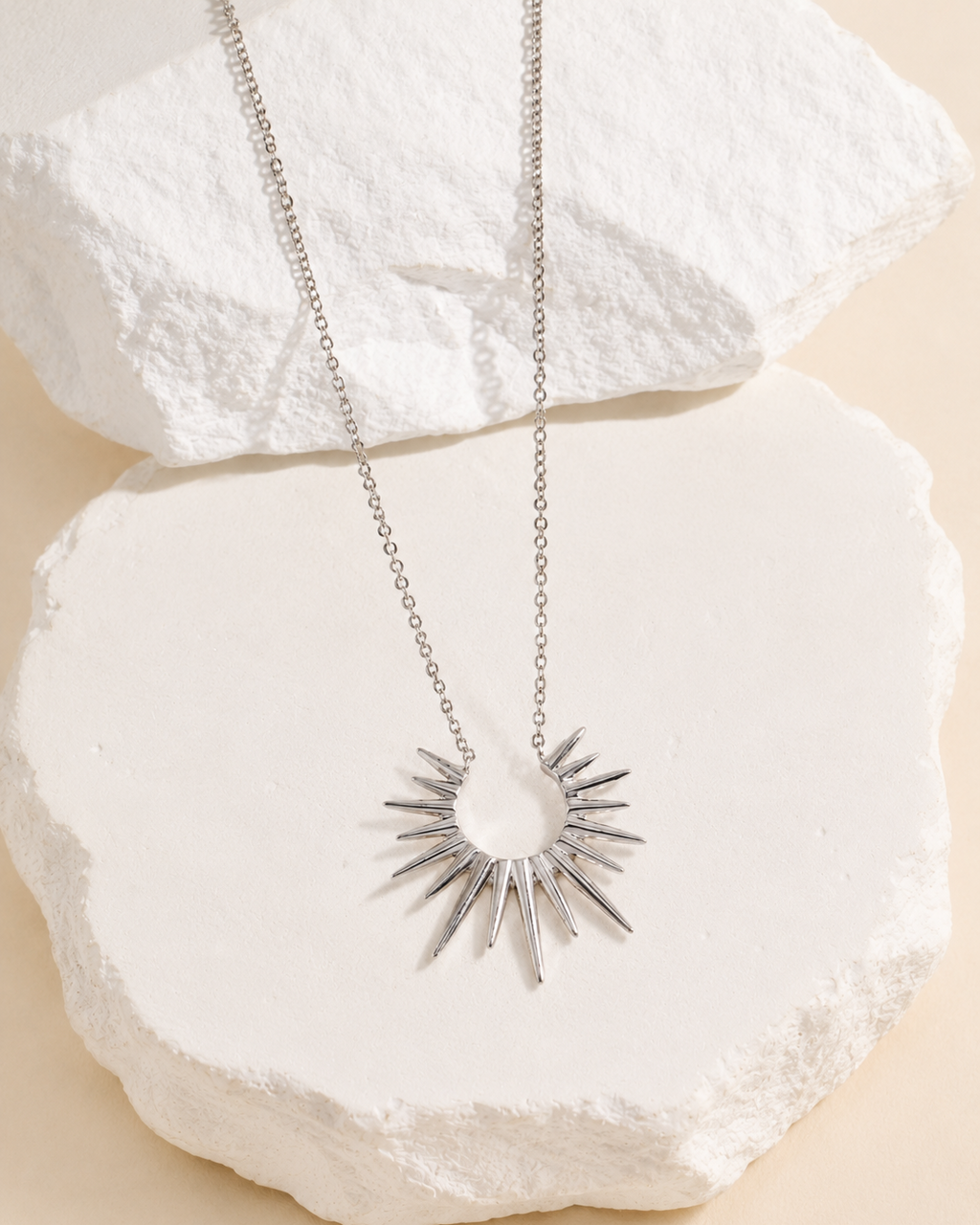 Silver sunburst pendant necklace displayed on a white textured stone slab against a soft beige background, featuring a radiant celestial sun design and fine chain.