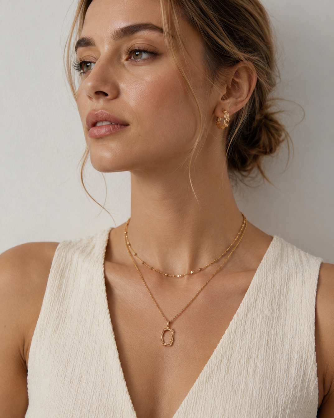European female model wearing a gold plated sculptural frame pendant necklace layered with a delicate beaded chain, styled with a cream textured dress.