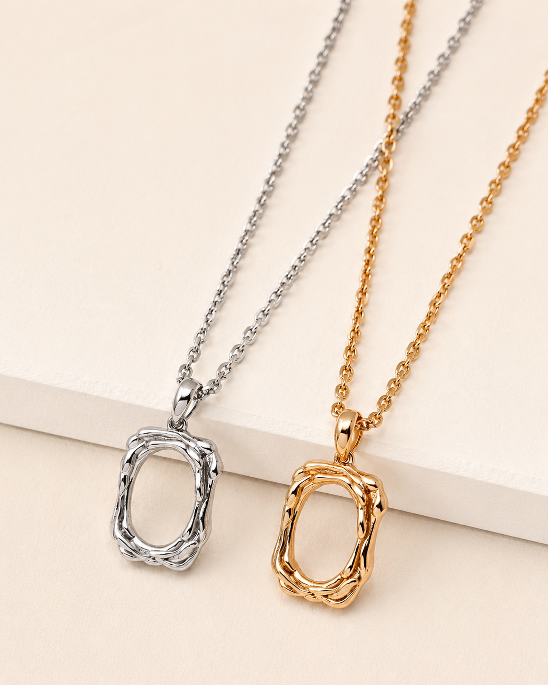 Gold and white gold plated sculptural frame pendant necklaces displayed side by side on a neutral beige surface, showcasing both color options.
