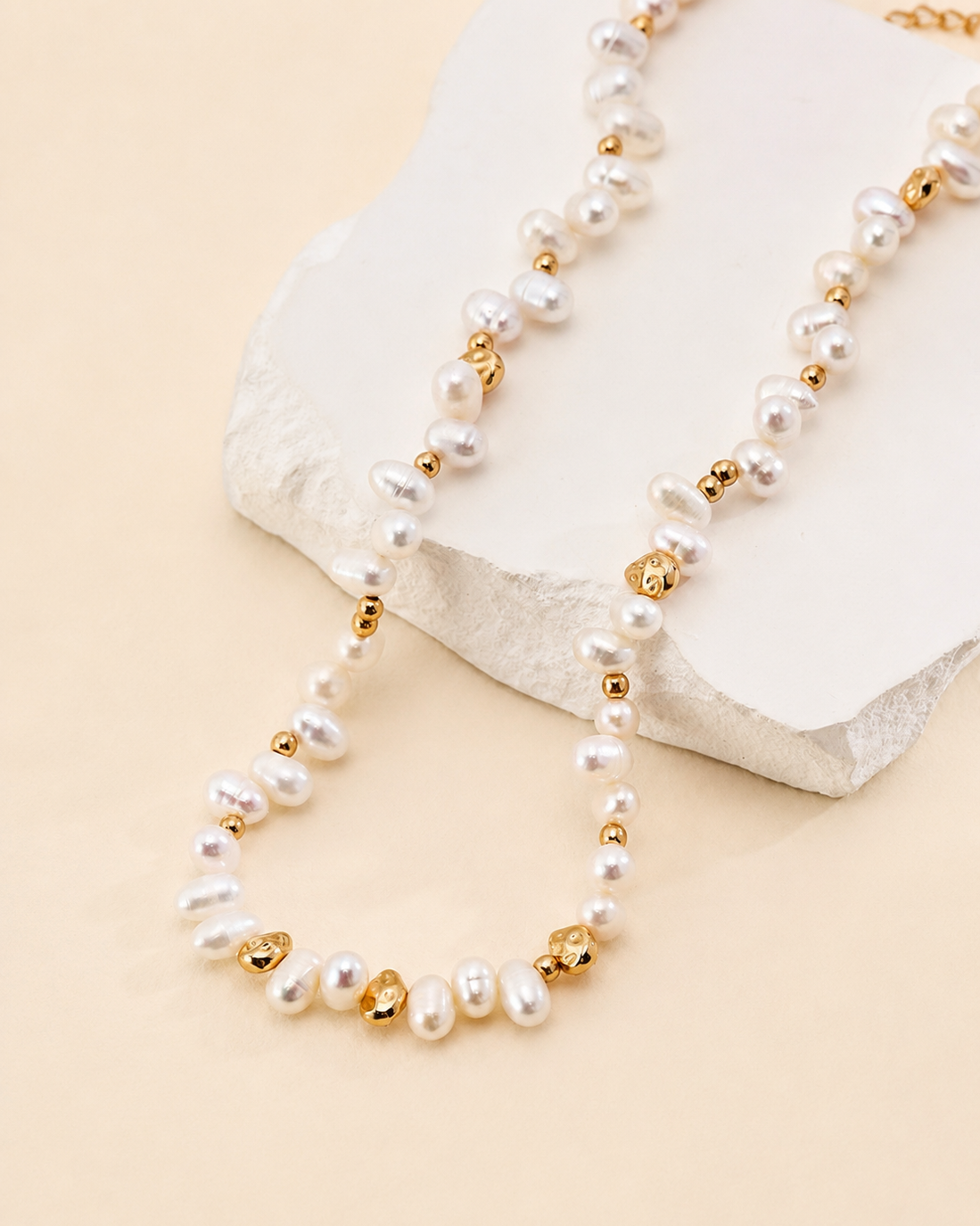 Freshwater pearl necklace with sculptural gold accents draped over a textured white stone pedestal on a soft beige background, elegant handcrafted jewelry.