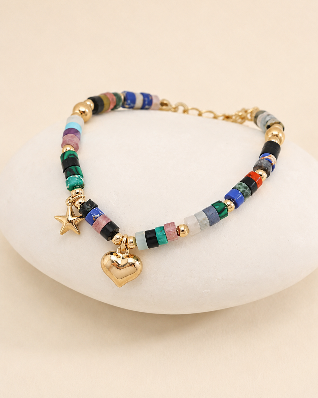 Close-up of a colorful natural gemstone bracelet featuring gold heart and star charms, displayed on a white stone pedestal.