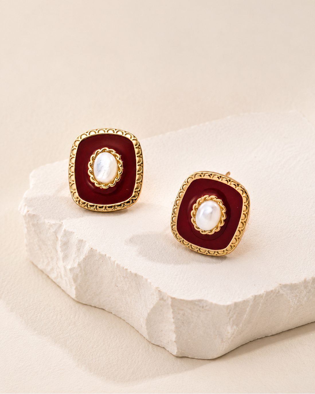 Red enamel and natural shell stud earrings with ornate gold-tone frame displayed on a textured white stone pedestal against a cream backdrop.