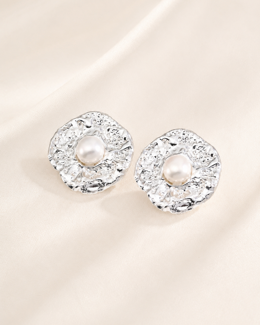 Silver textured stud earrings featuring centered pearls displayed on an elegant ivory fabric background.