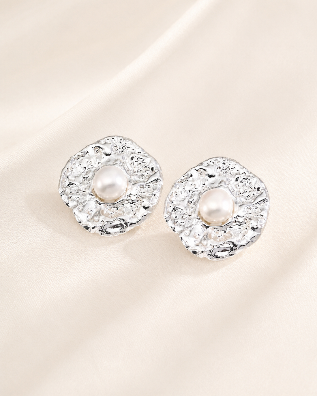 Silver textured stud earrings featuring centered pearls displayed on an elegant ivory fabric background.